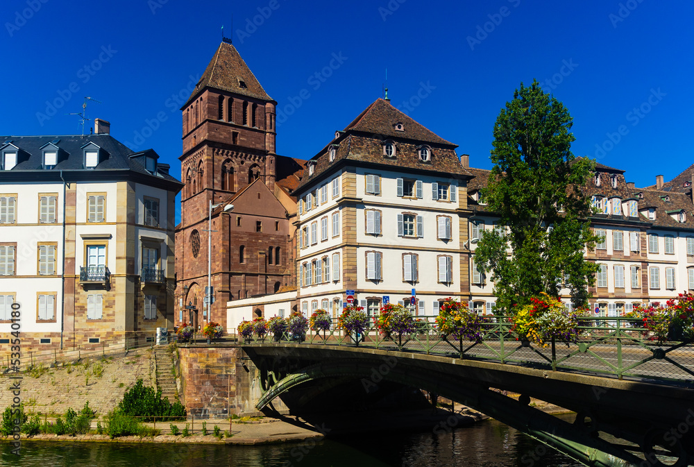 Obraz premium Scenic view of Strasbourg cityscape on sunny summer day overlooking historical building of St Thomas Church on bank of Ill river and bridge decorated with blooming flowers