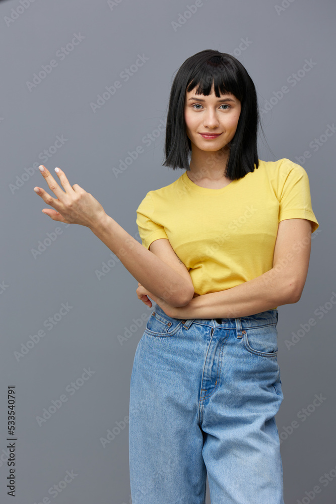 a happy, beautiful, attractive woman stands in a yellow T-shirt and light jeans on a gray background, posing with a relaxed hand gesturing. Vertical Studio Photography a happy, beautiful, attractive woman stands in a yellow T-shirt and light jeans on a gray background, posing with a relaxed hand gesturing. Vertical Studio Photography