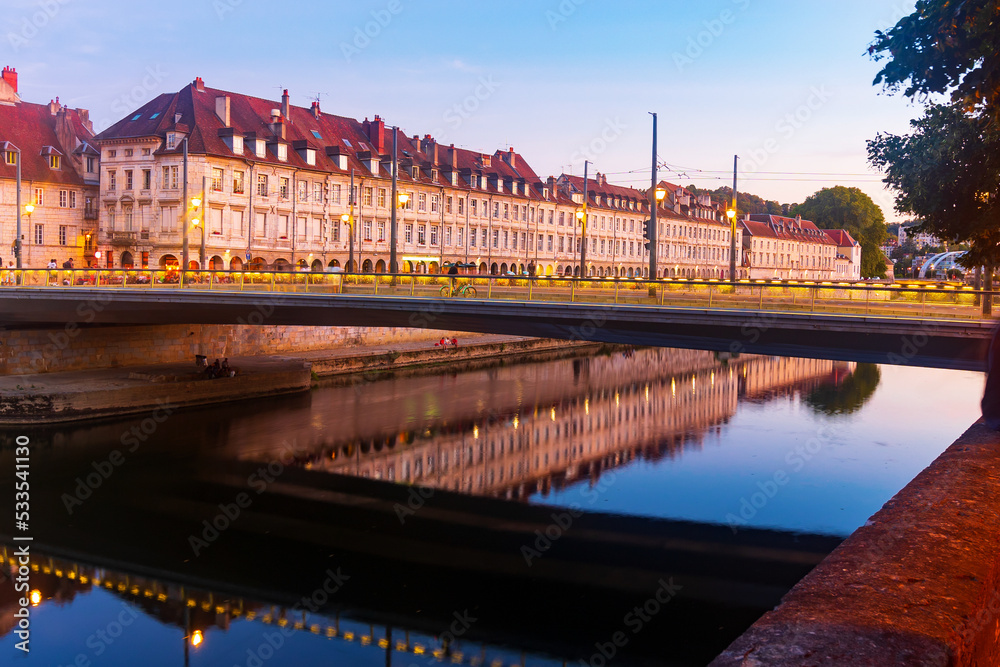 Picturesque view of architectural ensemble of illuminated embankment of Besancon and Battant bridge over Doubs river at dusk, France.