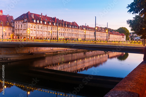 Picturesque view of architectural ensemble of illuminated embankment of Besancon and Battant bridge over Doubs river at dusk, France.