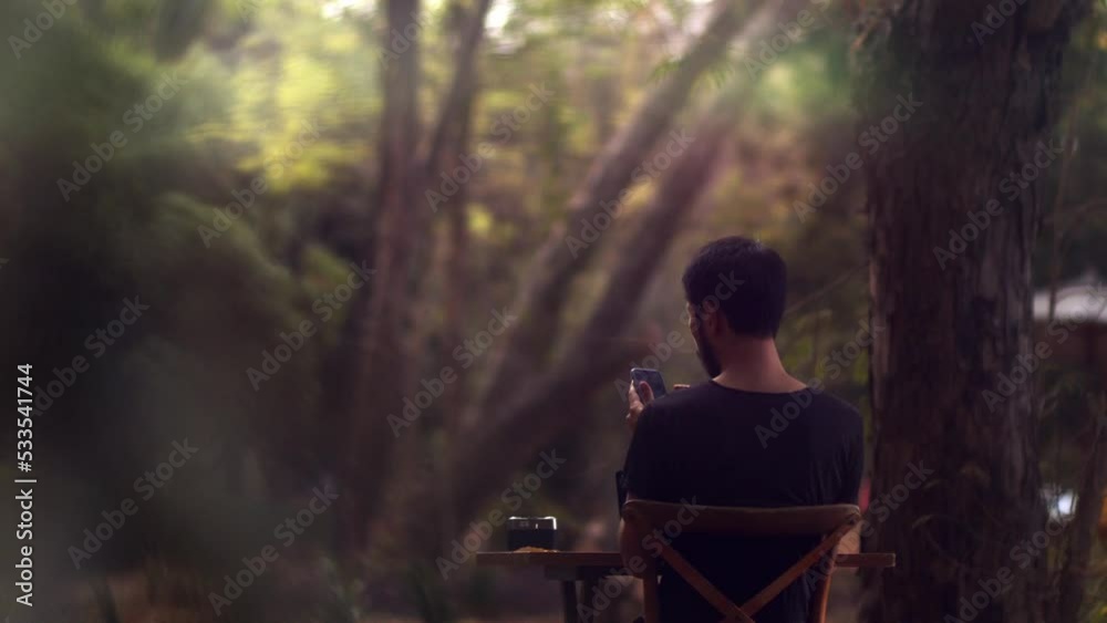 male model sitting in the garden at a table next to a tree checking messages on his smartphone - cinematic shot male model sitting in the garden at a table next to a tree checking messages on his smartphone - cinematic shot