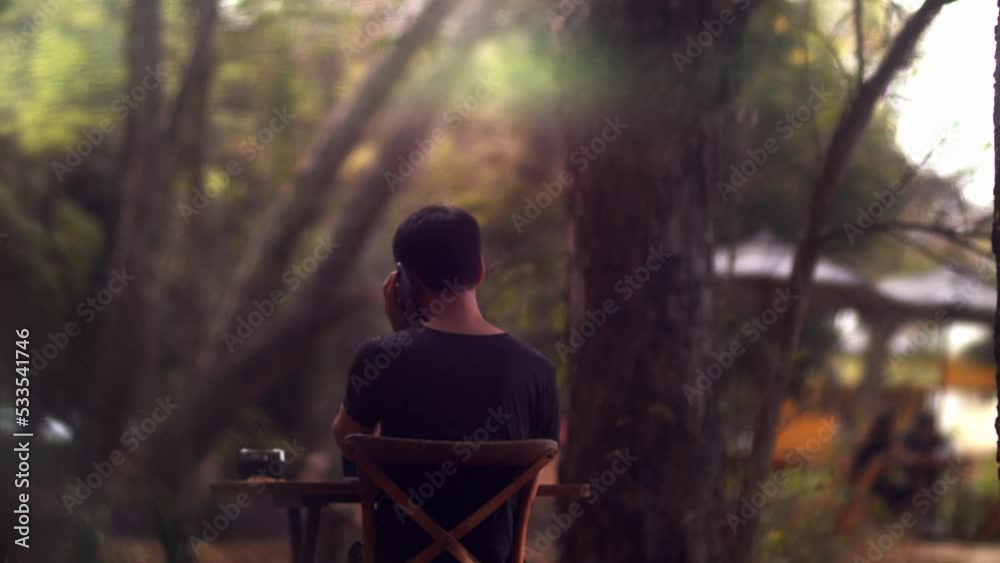 cinematic shot of a male model sitting at a table in the garden and taking his mobile phone for a call cinematic shot of a male model sitting at a table in the garden and taking his mobile phone for a call