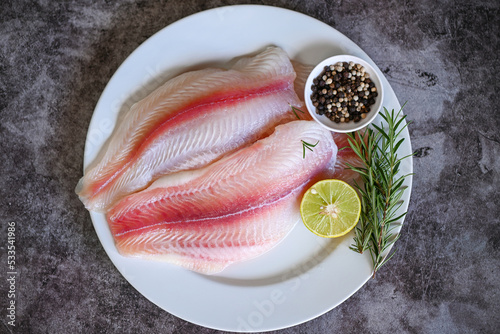 fresh raw pangasius fish fillet with herb and spices lemon lime and rosemary, meat dolly fish tilapia striped catfish, fish fillet on white plate with ingredients for cooking - top view photo