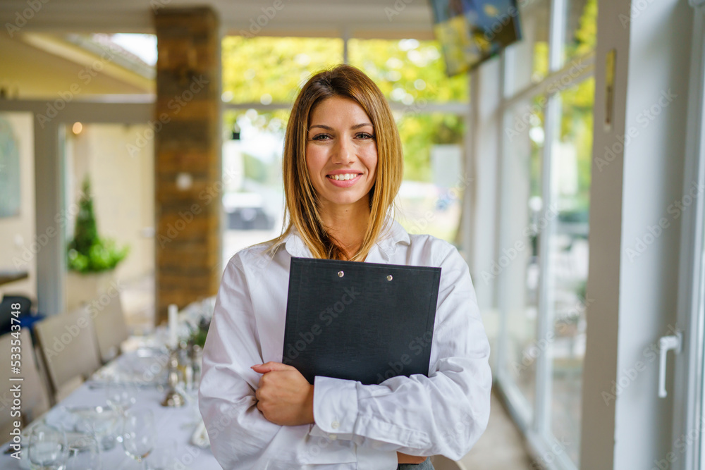 One woman young adult caucasian standing and smiling in the restaurant or hotel with paperwork in her hands and waiting clients Entrepreneur manager business modern lifestyle concept One woman young adult caucasian standing and smiling in the restaurant or hotel with paperwork in her hands and waiting clients Entrepreneur manager business modern lifestyle concept
