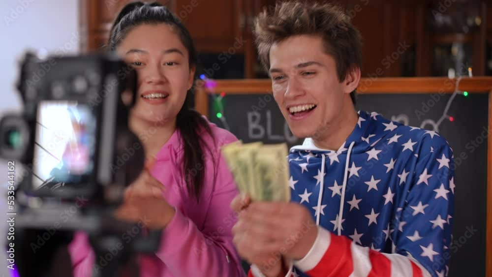 Grateful bloggers filming video on camera talking thanking subscribers for donations. Positive smiling Caucasian boy and Asian girl showing cash money sending air kisses recording vlog indoors