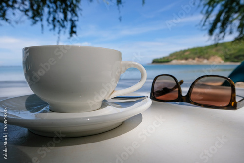 A white coffee cup and saucer and pair of sunglasses at a beachside cafe overlooking the ocean and blue sky on remote tropical island of Timor Leste