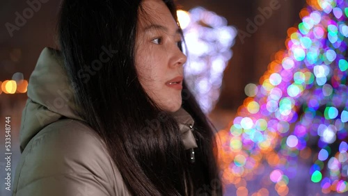 Side view close-up freezed Asian girl blowing on hands standing on Christmas evening outdoors. Teenager waiting on urban city street on New Year's eve with illumination light at background