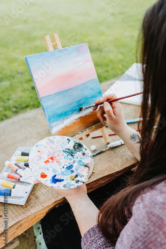 Fototapeta Naklejka Na Ścianę i Meble -  Unrecognizable young female artist painting her own beach scenery with oil on a canvas outdoor. Amateur painter.