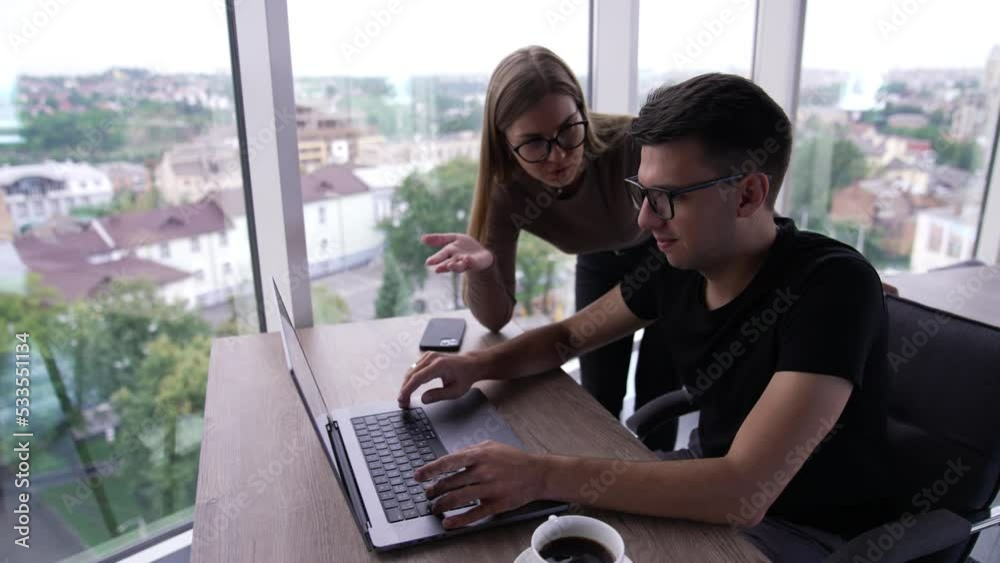 Colleagues look at the laptop screen point at it and discuss. Cooperative teammates solving work issues and brainstorming. Cityscape in window. Colleagues look at the laptop screen point at it and discuss. Cooperative teammates solving work issues and brainstorming. Cityscape in window.