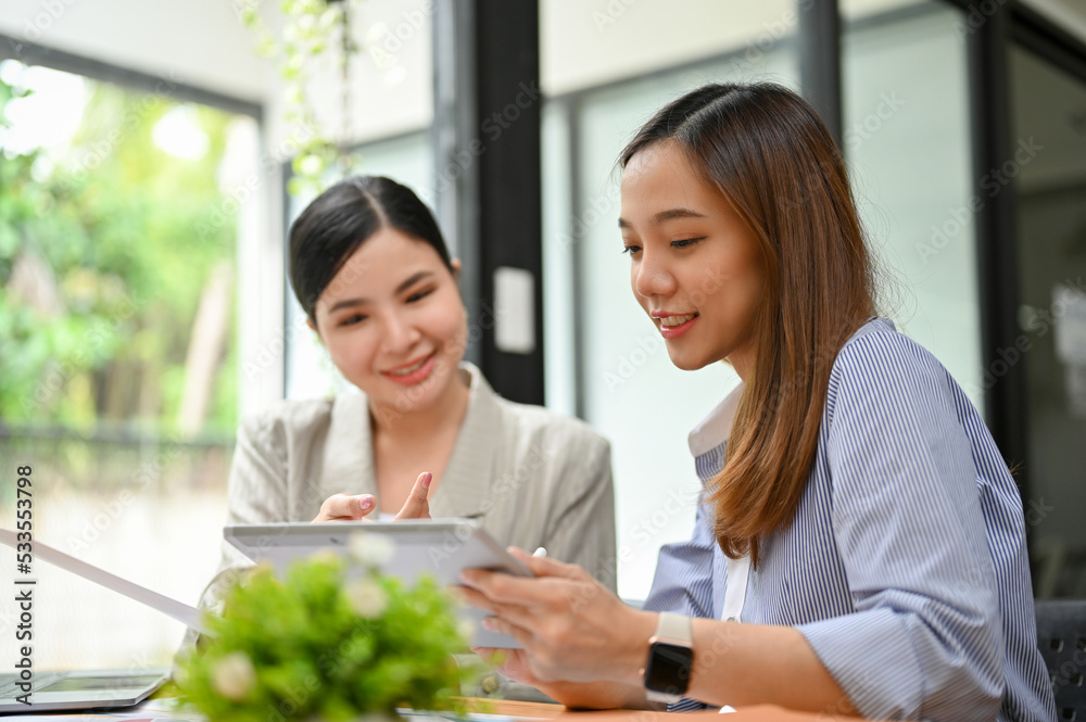 Charming millennial Asian businesswoman showing something on tablet screen to her colleague. Charming millennial Asian businesswoman showing something on tablet screen to her colleague.
