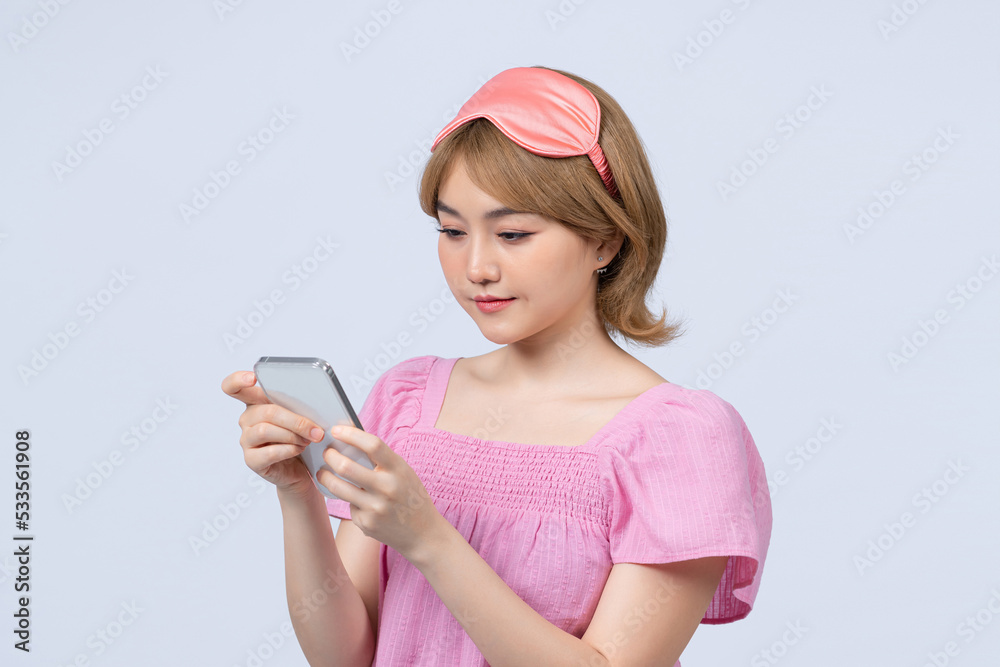 Happy young woman wears sleepmask uses mobile phone for sending text messages over gray background Happy young woman wears sleepmask uses mobile phone for sending text messages over gray background