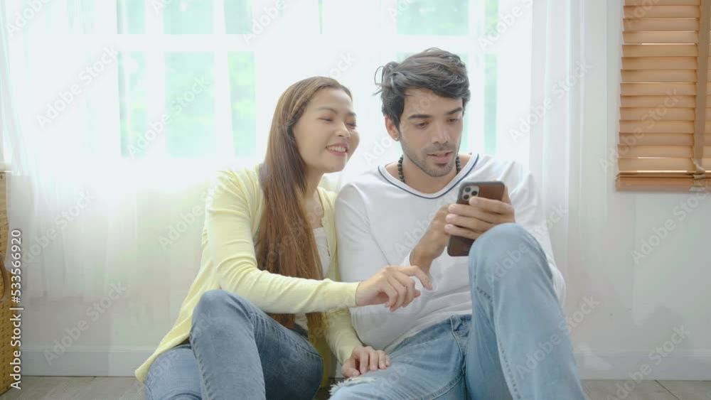 Relaxed young interracial couple or friends using a generic mobile phone together sitting in the living room at home, Happy young man and woman with smartphone, tease each other Relaxed young interracial couple or friends using a generic mobile phone together sitting in the living room at home, Happy young man and woman with smartphone, tease each other