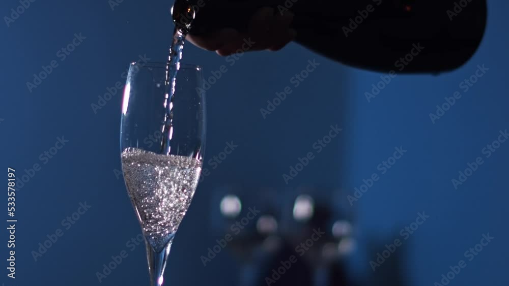 Champagne is poured from bottle into glass. Bubbles and foam rise in drink glass. Crystal glass with champagne.  Luxury white wine with bubbles. in slow motion in a blue light background