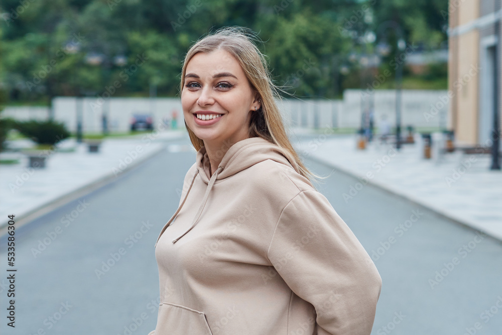 Young adult girl in a beige sweatshirt smiling, looking at the camera on the background of the urban environment Young adult girl in a beige sweatshirt smiling, looking at the camera on the background of the urban environment