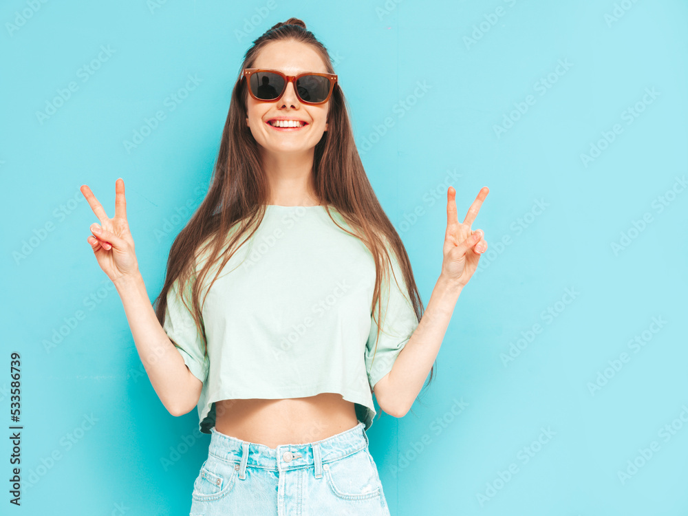 Young beautiful smiling female in trendy summer jeans skirt. carefree woman posing near blue wall in studio. Positive model having fun indoors in sunglasses. Cheerful and happy. Shows peace sign Young beautiful smiling female in trendy summer jeans skirt. carefree woman posing near blue wall in studio. Positive model having fun indoors in sunglasses. Cheerful and happy. Shows peace sign