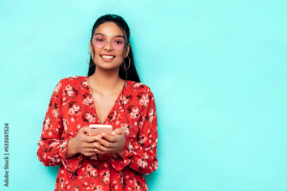 Young beautiful smiling female in trendy summer dress clothes. Sexy carefree woman posing near blue wall in studio. Positive model having using phone apps. Holding smartphone, looking at screen Young beautiful smiling female in trendy summer dress clothes. Sexy carefree woman posing near blue wall in studio. Positive model having using phone apps. Holding smartphone, looking at screen