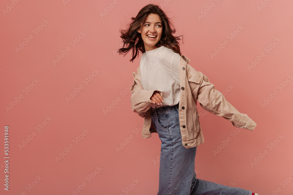 Active young caucasian brunette girl smiles looking away posing against pink background. Model with wavy hair wears T-shirt, jacket and jeans. Lifestyle concept is sincere people's emotions. Active young caucasian brunette girl smiles looking away posing against pink background. Model with wavy hair wears T-shirt, jacket and jeans. Lifestyle concept is sincere people's emotions.