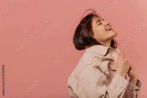 Joyful young caucasian girl smiles with mouth open, eyes closed on pink background with space for text. Model with dark hair wears beige jacket. Lifestyle concept