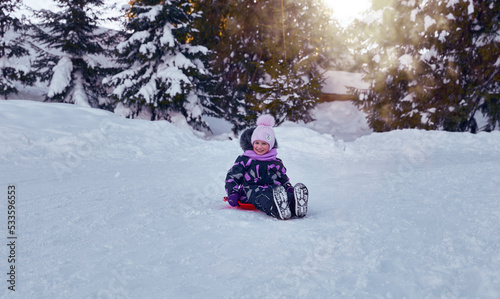 Little girl enjoying the winter sledding time. Child playing and having fun riding on a snowy hill. Slide down from snow slope sitting in plate