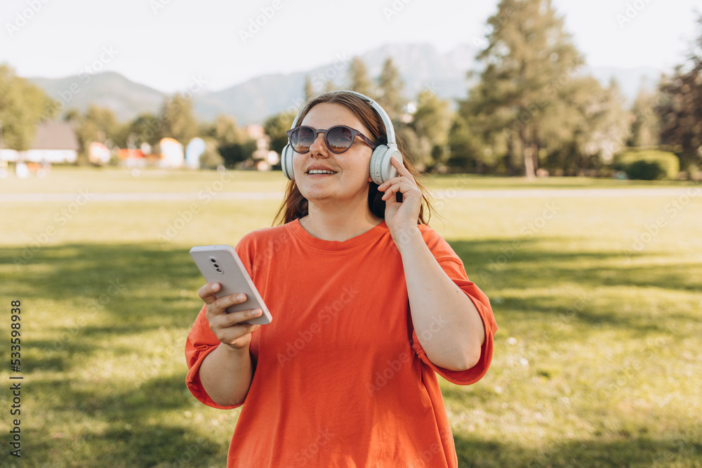 Close up portrait of a lovely happy young woman in sunglasses listening to music through wireless earphones on nature background. Music lover enjoying music. Gigl choosing sound track on cellphone Close up portrait of a lovely happy young woman in sunglasses listening to music through wireless earphones on nature background. Music lover enjoying music. Gigl choosing sound track on cellphone