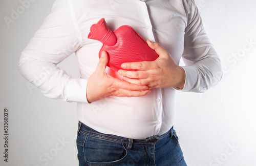 A man in a white shirt holds a red heating pad with hot water near the chest of the ribs. Treatment of intercostal neuralgia and pinched nerve, close-up photo