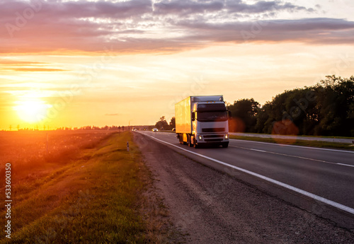 A truck with a trailer transports cargo in the summer against the backdrop of sunset and beautiful evening nature. The concept of logistics problems in trucking,  © HENADZY