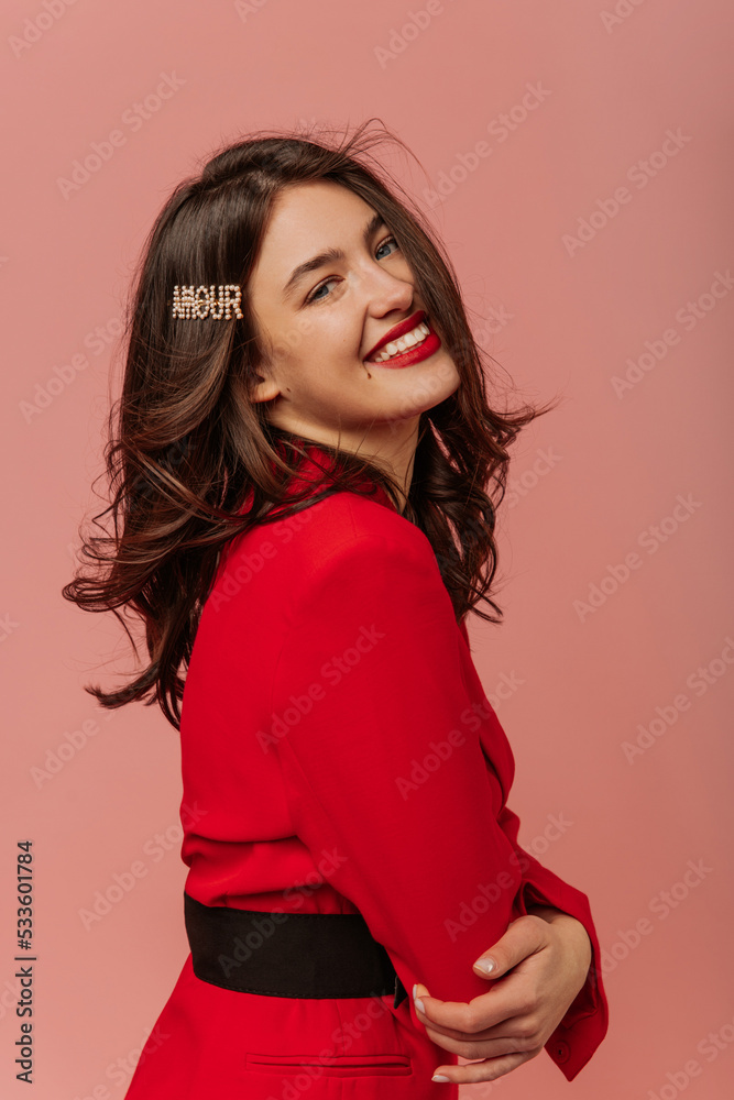 Happy young caucasian brunette girl smiles with her teeth at camera on pink background. Model with wavy hair is wearing red jacket. Mood, lifestyle, concept . Happy young caucasian brunette girl smiles with her teeth at camera on pink background. Model with wavy hair is wearing red jacket. Mood, lifestyle, concept .