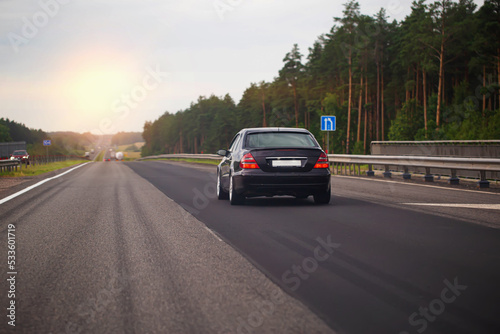 A black passenger car drives along an asphalt highway in the evening against the background of a sunset. The concept of traffic on the road and travel in your car. Copy space for text