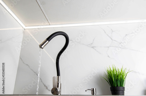 Contemporary kitchen faucet with curved spout set against white porcelain stoneware tiles. Water tap with black bending hose, close-up