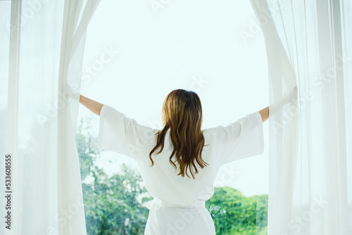 Back view of woman opening curtains and looking through the window in the summer within the bedroom at home.