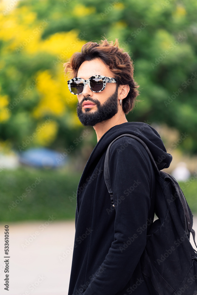 portrait of a beautiful young man with backpack and fashionable sunglasses walking by a city park, concept of urban lifestyle and stylish clothing, copy space for text portrait of a beautiful young man with backpack and fashionable sunglasses walking by a city park, concept of urban lifestyle and stylish clothing, copy space for text
