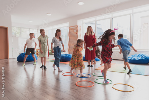 Fototapeta Naklejka Na Ścianę i Meble -  Small nursery school children with female teacher on floor indoors in classroom, doing exercise. Jumping over hula hoop circles track on the floor.
