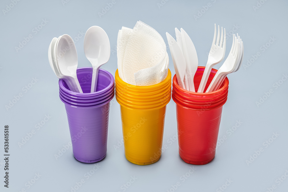 A set, in a row, of multi-colored disposable cups for drinks and cutlery. Grey background. A set, in a row, of multi-colored disposable cups for drinks and cutlery. Grey background.