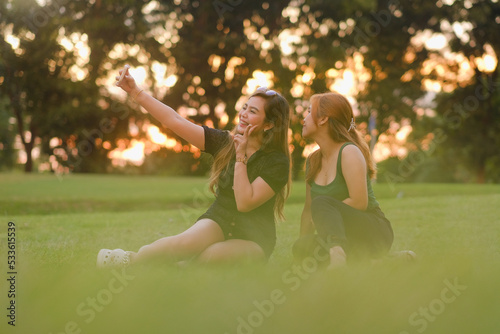 Two young Asian women pose for the camera as they take a selfie together. Both are sat on the grass in a park on a beautiful summer day at sunset. photo