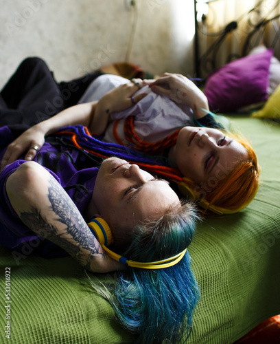 Portrait of a young tattooed punk couple of a girl and a guy with long dyed hair braided, lying on a large green bed, top view. They listen to music. Yellow large headphones. Stylish modern youth
