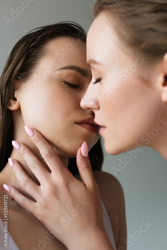 side view of young lesbian woman touching and kissing brunette girlfriend isolated on grey.