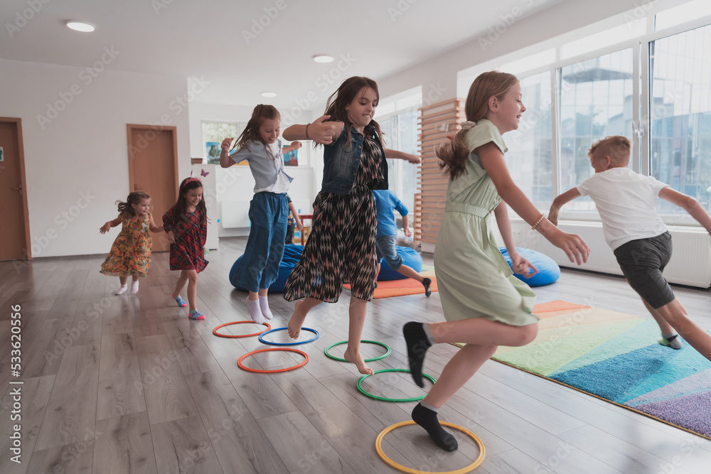 Fototapeta premium Small nursery school children with female teacher on floor indoors in classroom, doing exercise. Jumping over hula hoop circles track on the floor.