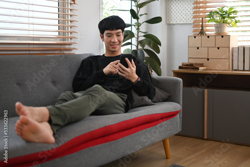 Man in warm sweater resting on couch and using mobile phone, enjoying leisure weekend time at home