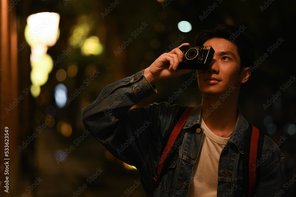Hipster man traveler taking photo with retro camera while standing in city with blurred night street lights background Hipster man traveler taking photo with retro camera while standing in city with blurred night street lights background