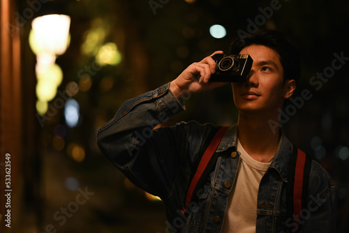 Hipster man traveler taking photo with retro camera while standing in city with blurred night street lights background © Prathankarnpap