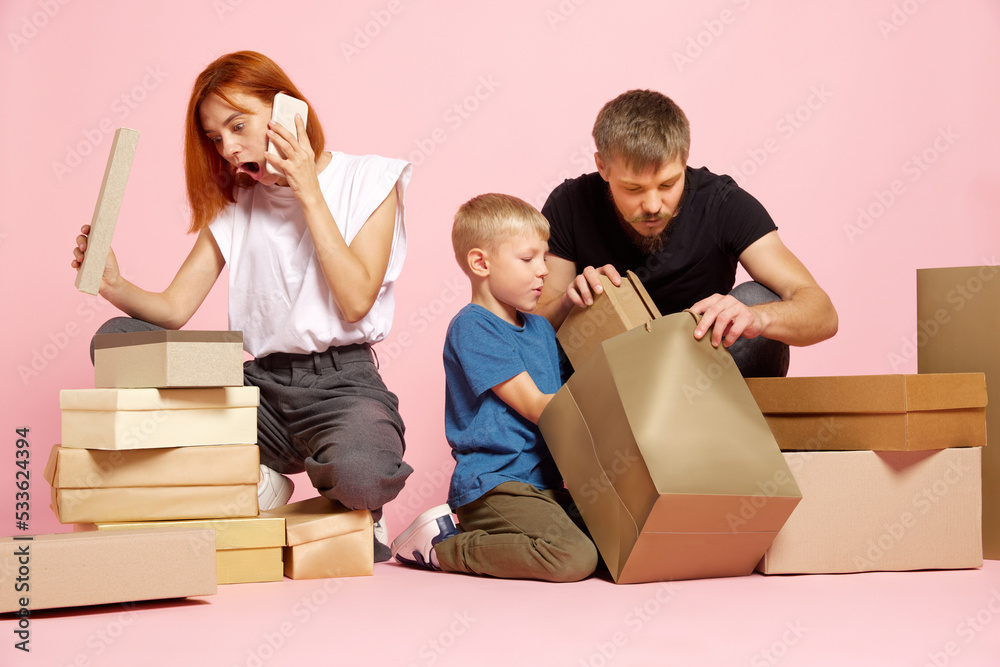 Wow. Happy mother, father and son open cardboard boxes, receive parcels unpack after online shopping over pink studio background. Seasonal sales concept
