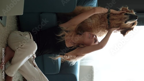 Young happy smiling blonde woman is caressing and kissing with affection her lovely labrador retriever dog while having fun together in living room at home. Vertical shoot. 4k footage