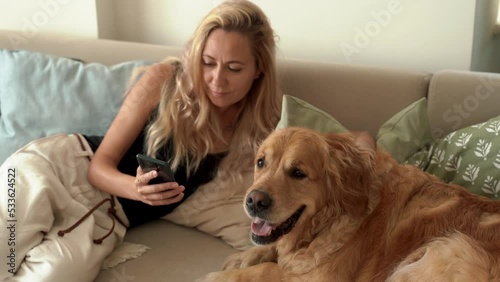 Young pretty woman using smartphone while sitting with her adorable golden retriever dog on the floor at cozy home. Domestic animal at home concept. 4k footage