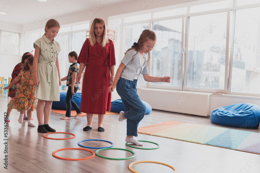 Fototapeta premium Small nursery school children with female teacher on floor indoors in classroom, doing exercise. Jumping over hula hoop circles track on the floor.