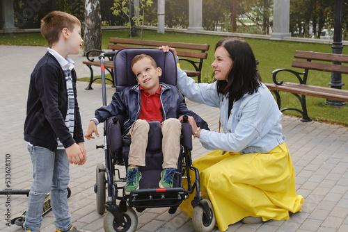 Family with cerebral palsy child on special wheelchair walking outdoors. Integration and accessibility of disabled people, inclusion. Brother with physical disorder Mother taking care of handicap kid © ninelutsk