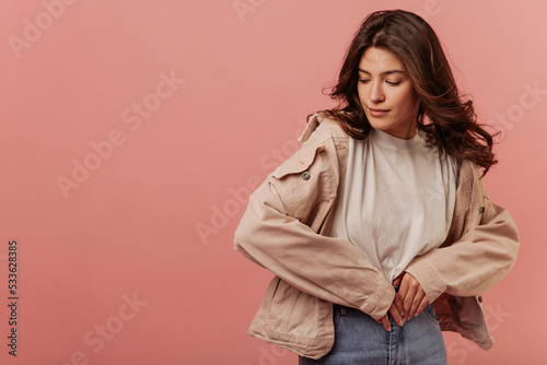 Modern young caucasian girl averts gaze holding hands to her belly on pink background. Brunette girl with wavy hair wears light-colored casual clothes. Lifestyle, leisure concept.  © Look!