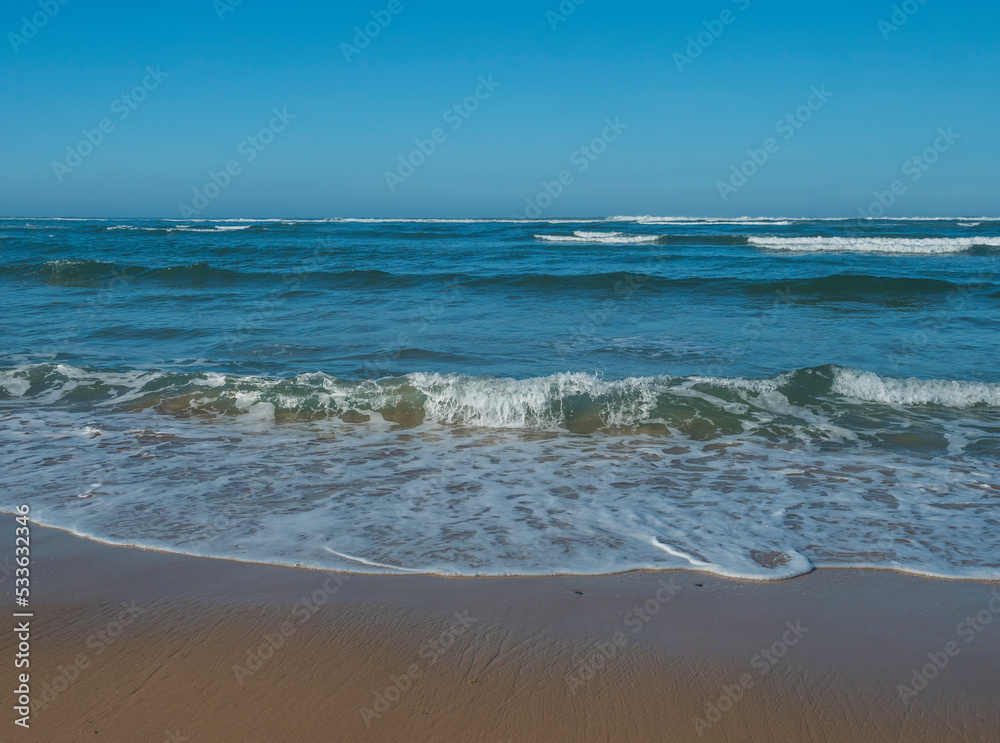 Scenic view turquoise blue breaking sea waves with white foam approaching the golden sand beach at wild Rota Vicentina coast at Portugal. Atlantic ocean vawes with blue sky at sunny autumn day
