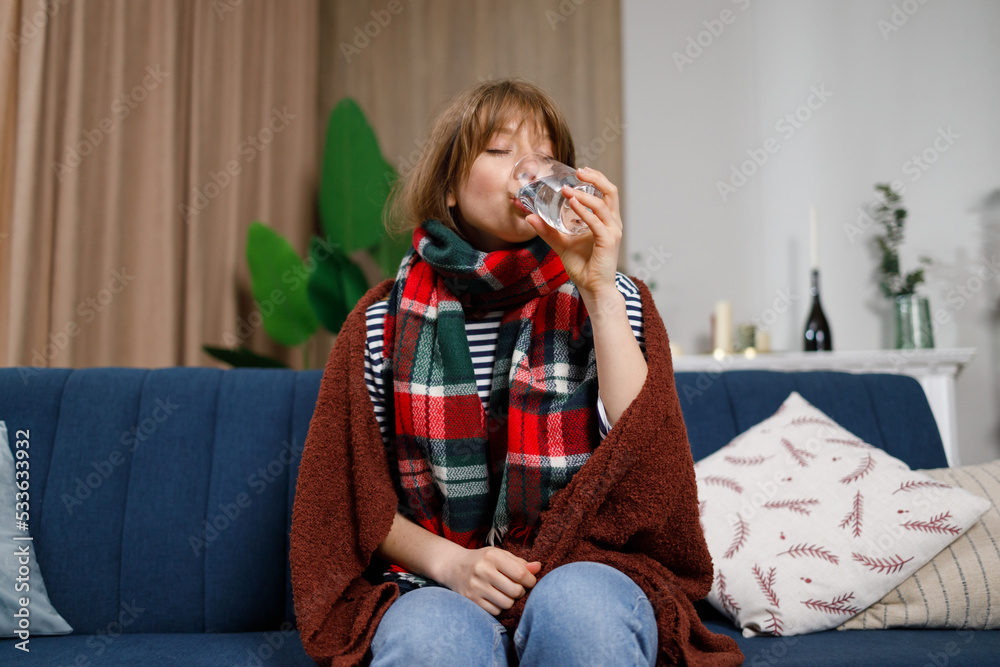 Sick girl drinks water sitting on the couch. Young woman being treated for flu at home