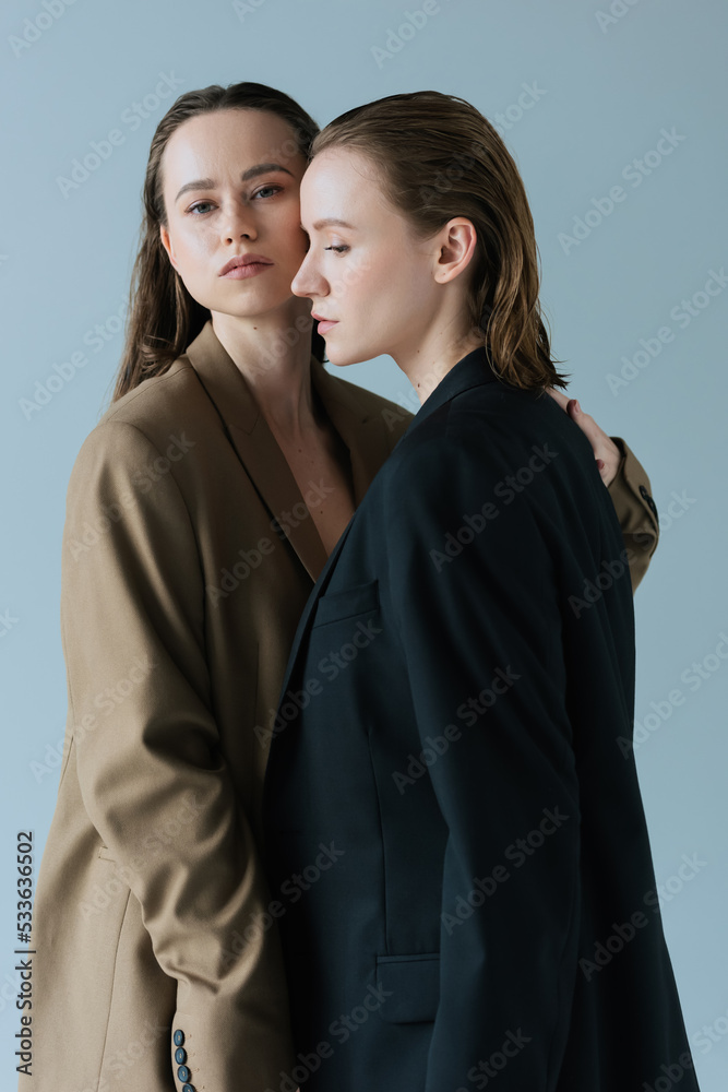 young woman in beige blazer hugging lesbian partner and looking at camera isolated on grey.