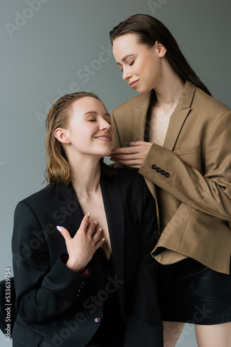 brunette woman touching face of happy girlfriend sitting with closed eyes isolated on grey.